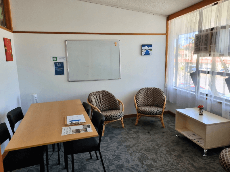 Full view of meeting room 5. Table and chairs to the left with single comfy chairs by the back wall. Window to the right with curtains open and natural light, whiteboard on the wall