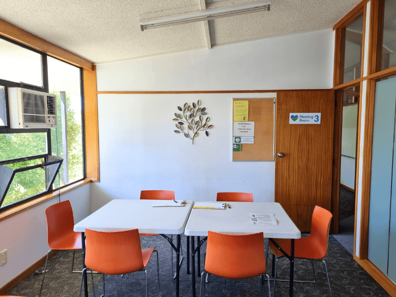 View of meeting room 3 from the back of the room with door open and sign on the door indicating room number. Decoration on the wall with table and orange chairs in them middle of the room